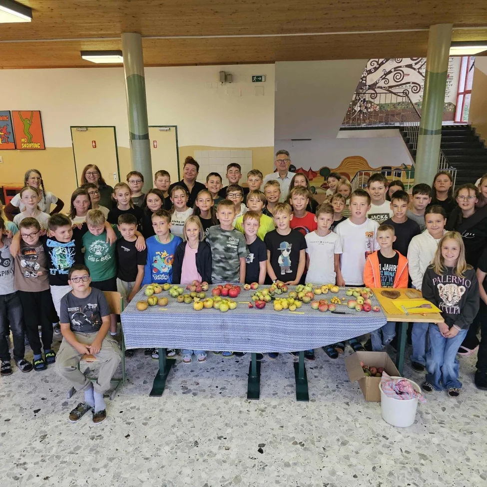Eine Gruppe von Kindern und Erwachsenen versammelt sich um einen Tisch voller Äpfel und posiert für ein Foto in einer Schulumgebung.