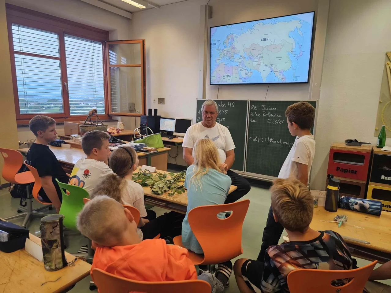 Ein Klassenzimmer mit einem Lehrer, der eine Weltkarte mit Schülern diskutiert. Der Lehrer steht vor einer Tafel mit Notizen. Schüler sitzen auf Stühlen, einige schauen den Lehrer an, während andere die Karte betrachten.