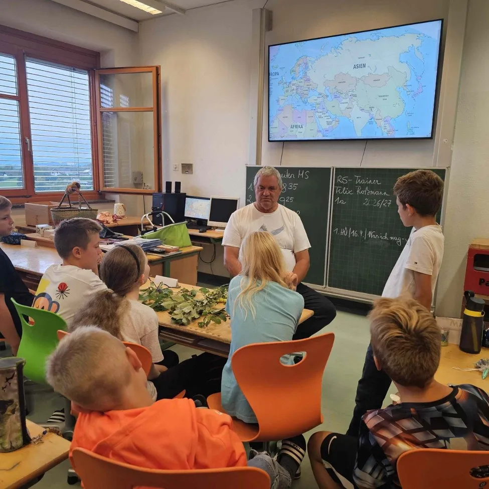 Ein Klassenzimmer mit einem Lehrer, der eine Weltkarte mit Schülern diskutiert. Der Lehrer steht vor einer Tafel mit Notizen. Schüler sitzen auf Stühlen, einige schauen den Lehrer an, während andere die Karte betrachten.