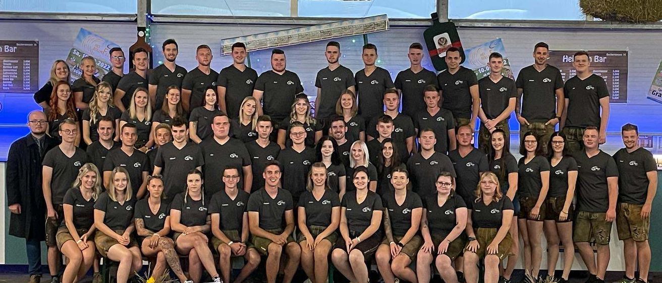 Eine große Gruppe von Menschen in schwarzen Shirts posiert für ein Foto unter einem Banner, das 'HeiBollm Bar' steht. Einige tragen Brillen, Armbänder und Turnschuhe. Es gibt einen Becher und einen Blumenvase auf der Bühne.