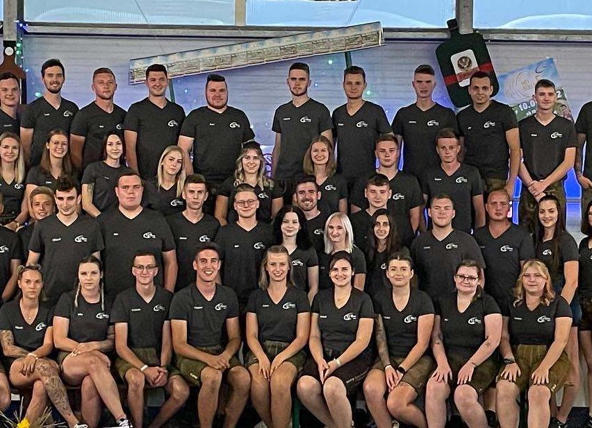 Eine große Gruppe von Menschen in schwarzen Shirts posiert für ein Foto unter einem Banner, das 'HeiBollm Bar' steht. Einige tragen Brillen, Armbänder und Turnschuhe. Es gibt einen Becher und einen Blumenvase auf der Bühne.