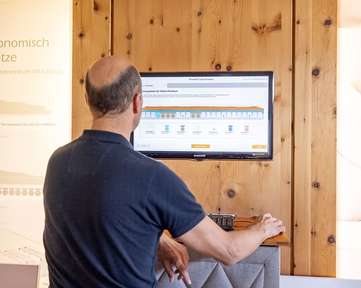 Ein kahlköpfiger Mann in einem blauen Shirt schaut auf einen Samsung-Monitor. Dieser zeigt einen Simulator für das Produkt Vitario Premium. Er steht vor einer Holzwand.