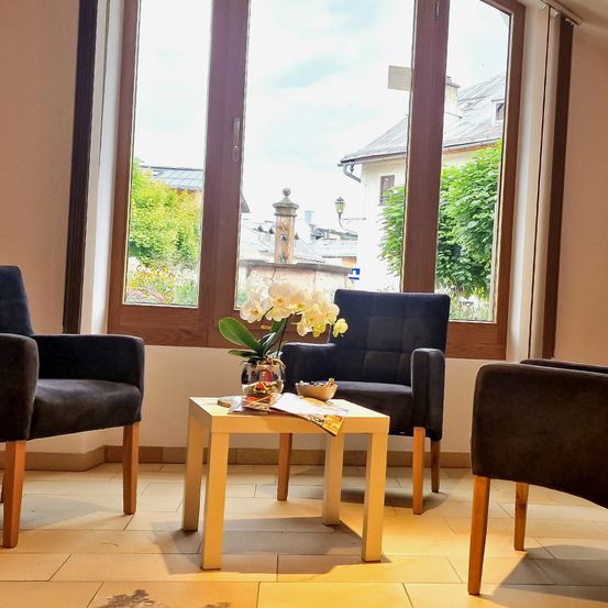 Ein Raum mit Holzstühlen und einem zentralen Tisch mit einem Blumenvase und einigen Büchern. Der Raum hat ein großes Fenster mit Blick auf ein Gebäude und Bäume.