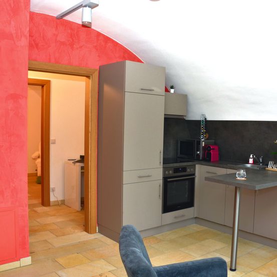 Eine moderne Küche mit roten Wänden, einem offenen Türeingang, einem Herd, einer Mikrowelle und einer Arbeitsplatte. Ein grauer Stuhl steht vor der Arbeitsplatte.