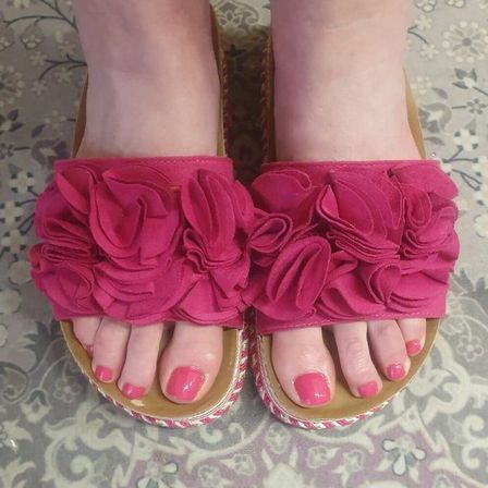 Ein Paar rosa Sandalen mit gerüschten Riemen, getragen von einer Person mit lackierten Zehennägeln.