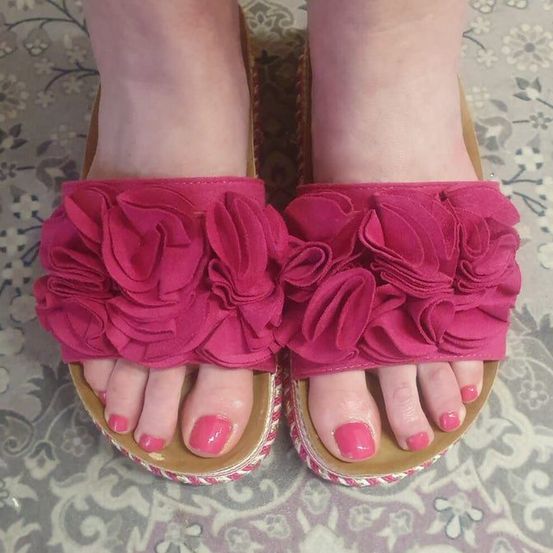 Ein Paar rosa Sandalen mit gerüschten Riemen, getragen von einer Person mit lackierten Zehennägeln.