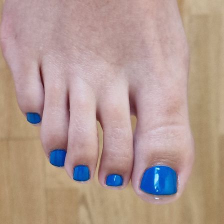 Ein Nahaufnahme eines Fußes mit in leuchtendem Blau lackierten Zehennägeln. Die Zehennägel sind ordentlich geschnitten und der Fuß ruht auf einer Holzoberfläche.