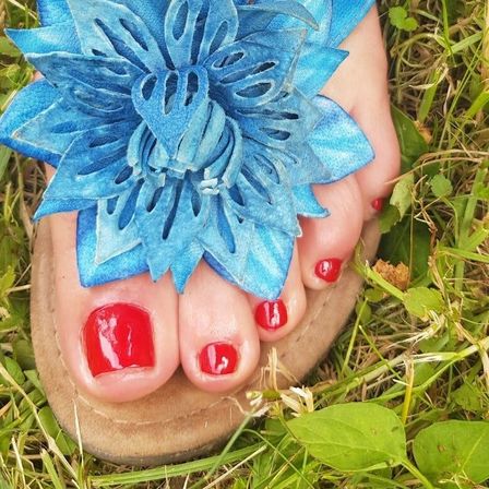 Ein Fuß mit leuchtend roten Nägeln, geschmückt mit einer blauen Blume, befindet sich auf einem Grasfeld.