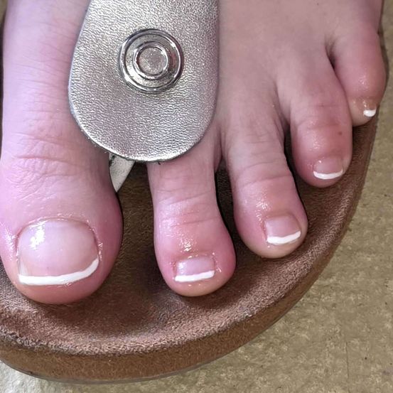 Ein Nahaufnahme von Füßen mit frisch lackierten Zehennägeln. Die Zehennägel haben eine weiße Basis mit einem hellen rosa Farbton. Der große Zeh wird mit einer silbernen Nagelschere geschnitten.