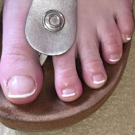 Ein Nahaufnahme von Füßen mit frisch lackierten Zehennägeln. Die Zehennägel haben eine weiße Basis mit einem hellen rosa Farbton. Der große Zeh wird mit einer silbernen Nagelschere geschnitten.