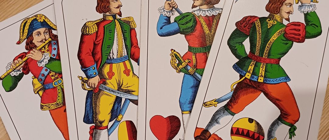 Vier Spielkarten liegen auf einer Holzoberfläche. Jede Karte zeigt einen Mann in einer anderen farbigen Uniform, der Waffen hält und heldenhaft posiert.