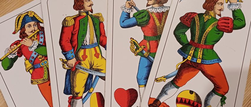 Vier Spielkarten liegen auf einer Holzoberfläche. Jede Karte zeigt einen Mann in einer anderen farbigen Uniform, der Waffen hält und heldenhaft posiert.
