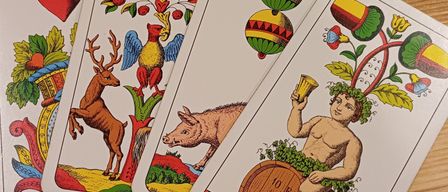 Vier Spielkarten sind auf einer Holzoberfläche ausgestellt. Die Karten zeigen verschiedene Illustrationen: ein Hirsch, ein Schwein, einen Ballon und einen Satyr, der auf einem Fass sitzt.
