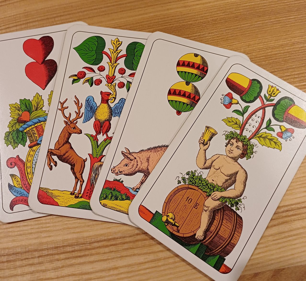 Four playing cards with various illustrations are on a wooden surface. One card has two hearts, another features a deer, a bird, and a tree. A third card depicts a pig, and the last card shows a boy sitting on a barrel.