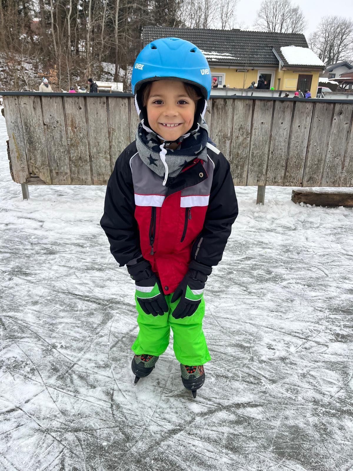 Ein junger Junge in Winterkleidung, einschließlich Helm und Schlittschuhen, steht auf einer Eisbahn. Hinter ihm befindet sich ein Holzzaun und ein Gebäude mit Menschen.