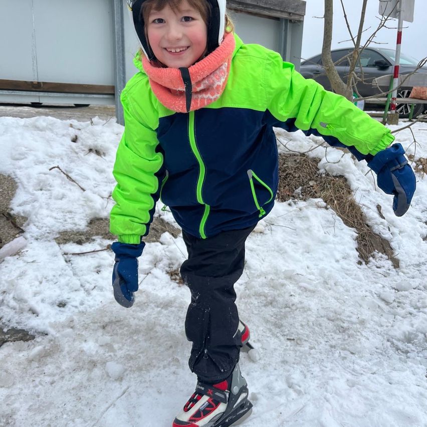 Ein junges Kind in Winterkleidung rutscht mit lächelndem Gesicht auf Schnee, ein Auto geparkt dahinter und ein Baum mit einem Pfosten in der Nähe.