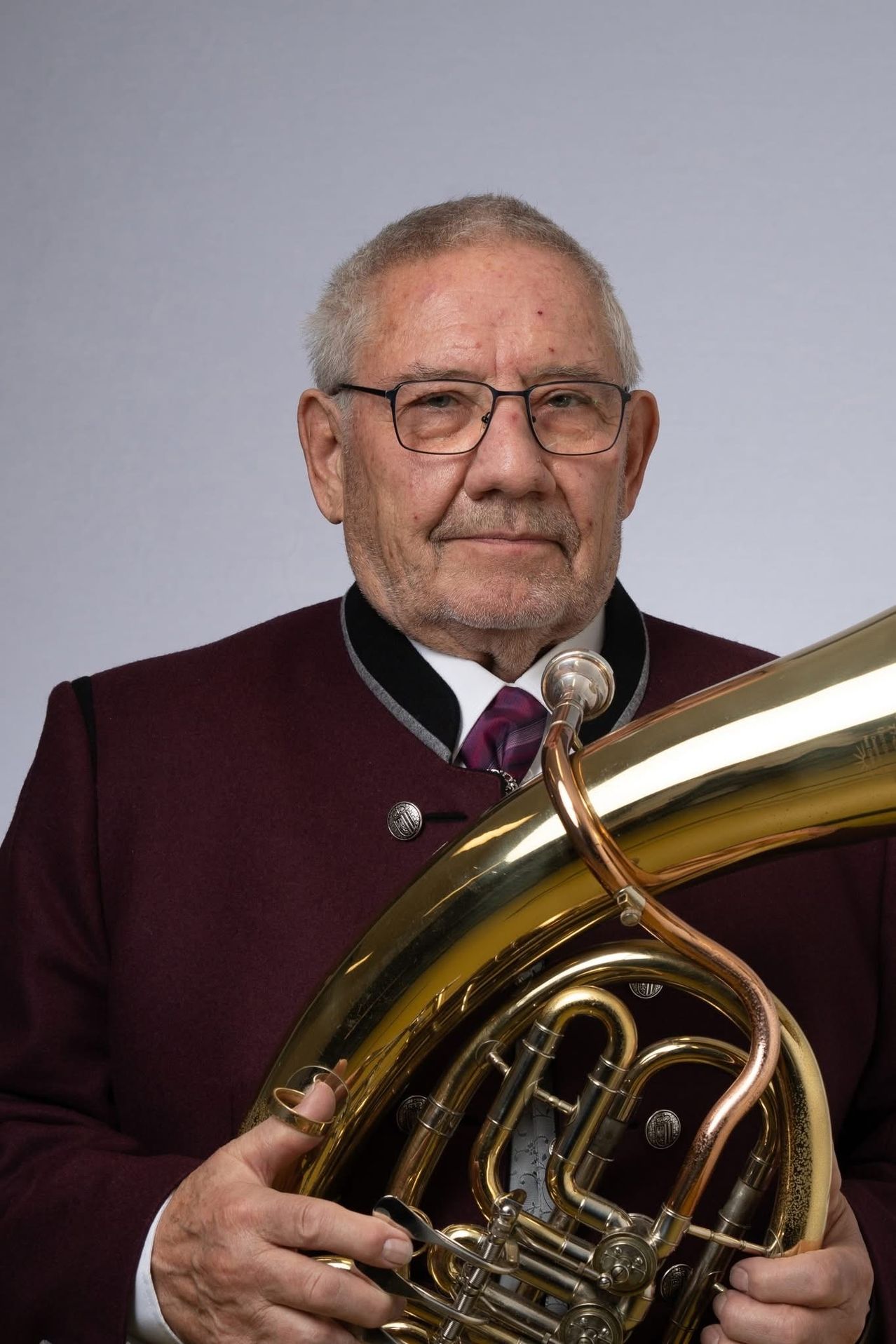 Ein älterer Mann lächelt, während er ein Blechinstrument hält. Er trägt einen dunkelroten Mantel, eine violette Krawatte und eine Brille.