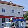 Ein weißer Lkw mit einem blauen Kran steht vor einem Gebäude mit dem Namen Elektro-Schweitzer. Das Gebäude hat ein rotes Dach und mehrere Fenster. Der Himmel ist klar.