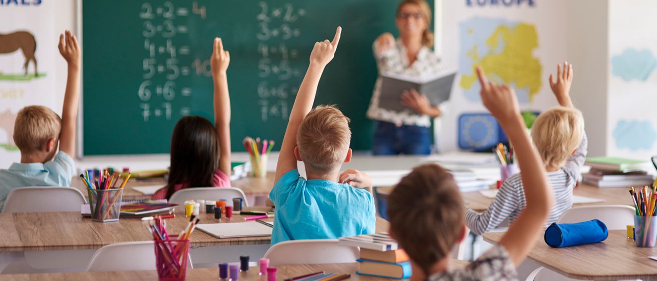 Ein Klassenzimmer mit Kindern, die ihre Hände heben, um eine Frage der Lehrerin zu beantworten. Die Lehrerin steht vor einer Tafel mit Matheproblemen.
