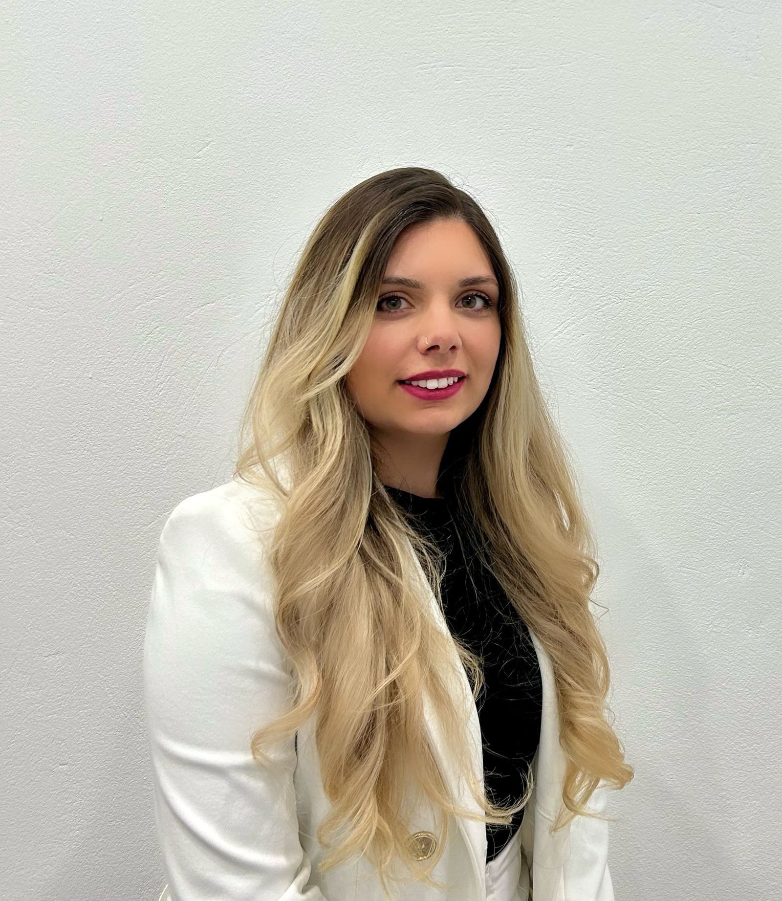 Eine Frau mit langen blonden Haaren lächelt für ein Foto. Sie trägt einen weißen Blazer über einem schwarzen Oberteil. Der Hintergrund ist eine schlichte, helle Wand.