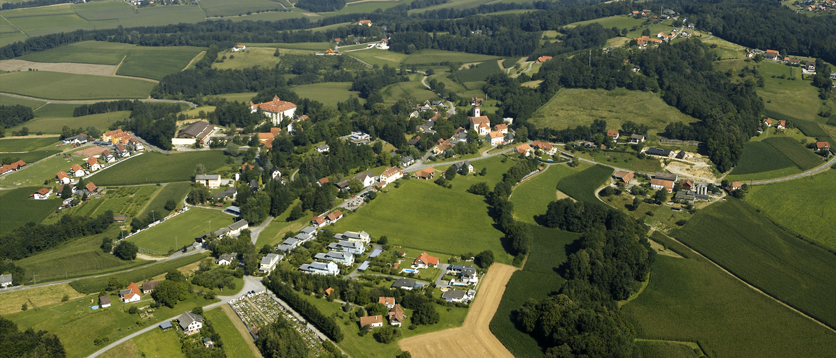 Ein Luftbild eines ländlichen Dorfes mit vielen Häusern und Bäumen, umgeben von grünen Feldern und landwirtschaftlichen Flächen, mit einem klaren blauen Himmel in der Ferne.