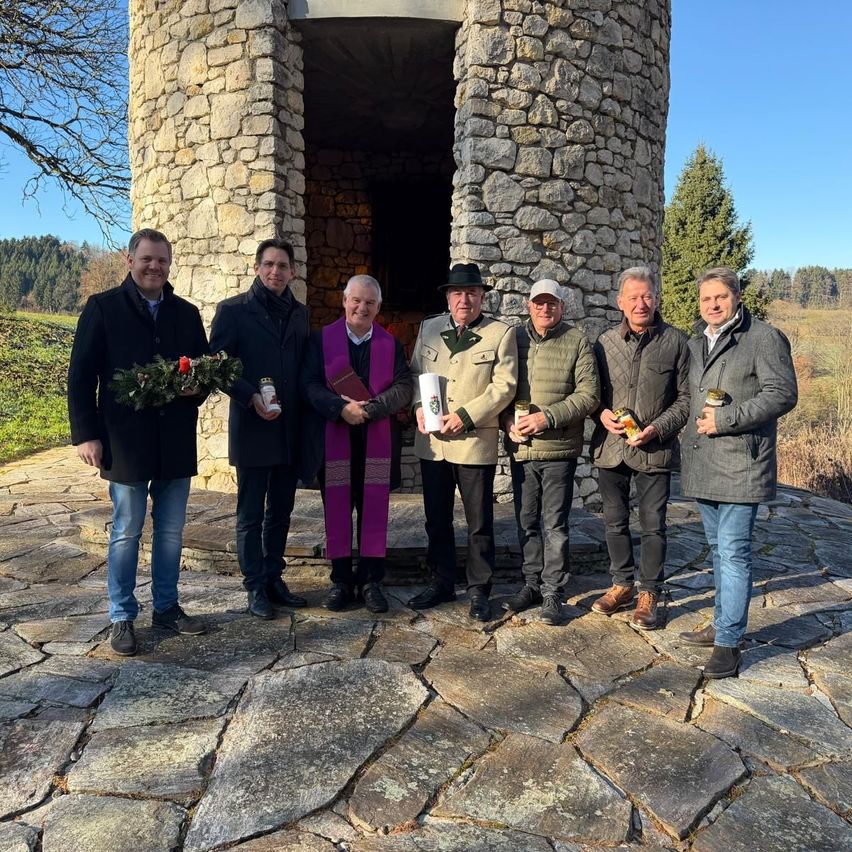 Eine Gruppe von Männern steht vor einem steinernen Gebäude. Einer hält einen Kranz, ein anderer ein Buch. Sie sind in Mänteln und Hosen gekleidet. Hinter ihnen ist eine Landschaft mit Bäumen und Büschen sichtbar.