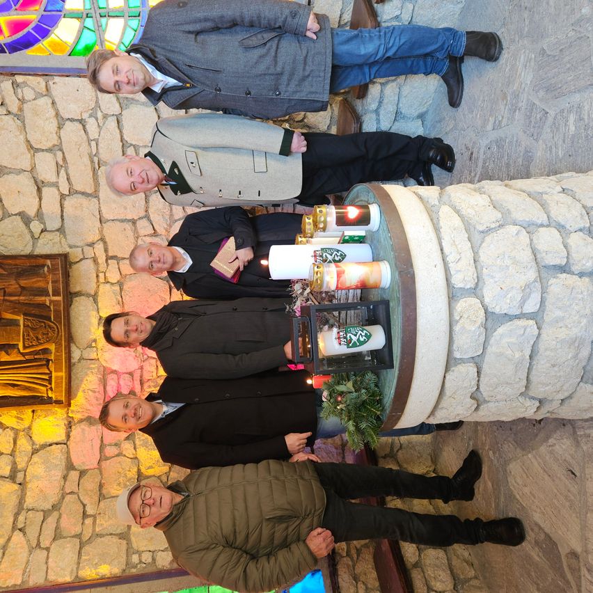 Fünf Männer posieren für ein Foto in einer Kirche. Vier stehen um ein Taufbecken mit Kerzen. Der Mann in der Mitte hält ein Buch. Die Wand hinter ihnen besteht aus Stein.