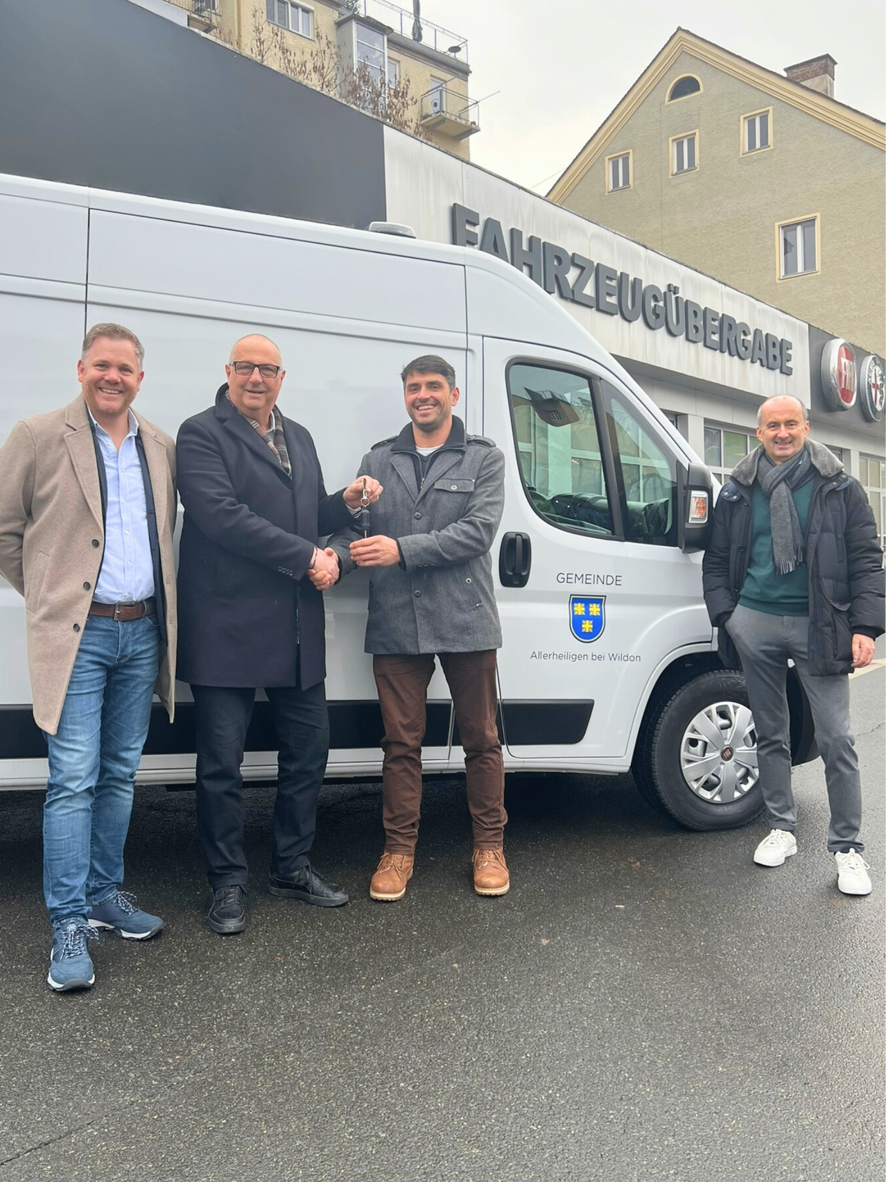 Vier Männer stehen vor einem weißen Van, zwei halten einen Schlüssel. Sie lächeln und schauen in die Kamera. Hinter ihnen befindet sich ein Gebäude mit einem Schild, das 'Fahrzeugübergabe' besagt.