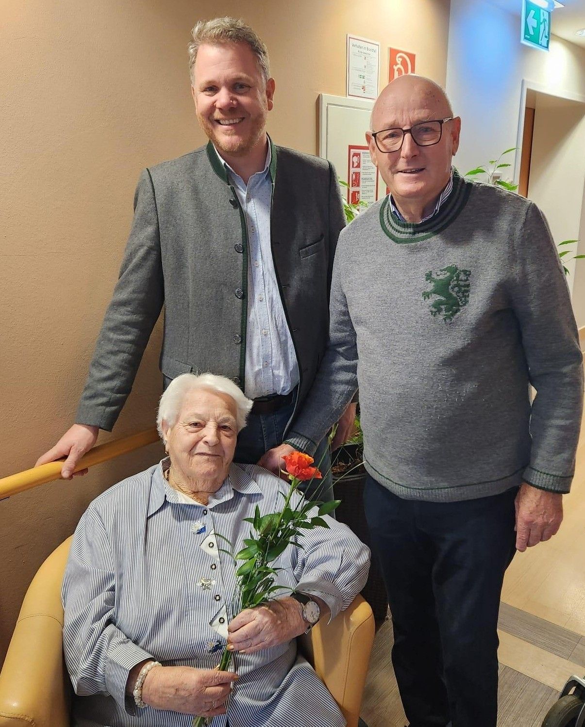 Drei Menschen stehen drinnen zusammen. Eine ältere Frau hält einen Blumenstrauß. Der Mann hinter ihr lächelt. Der Mann neben ihr trägt eine Brille.