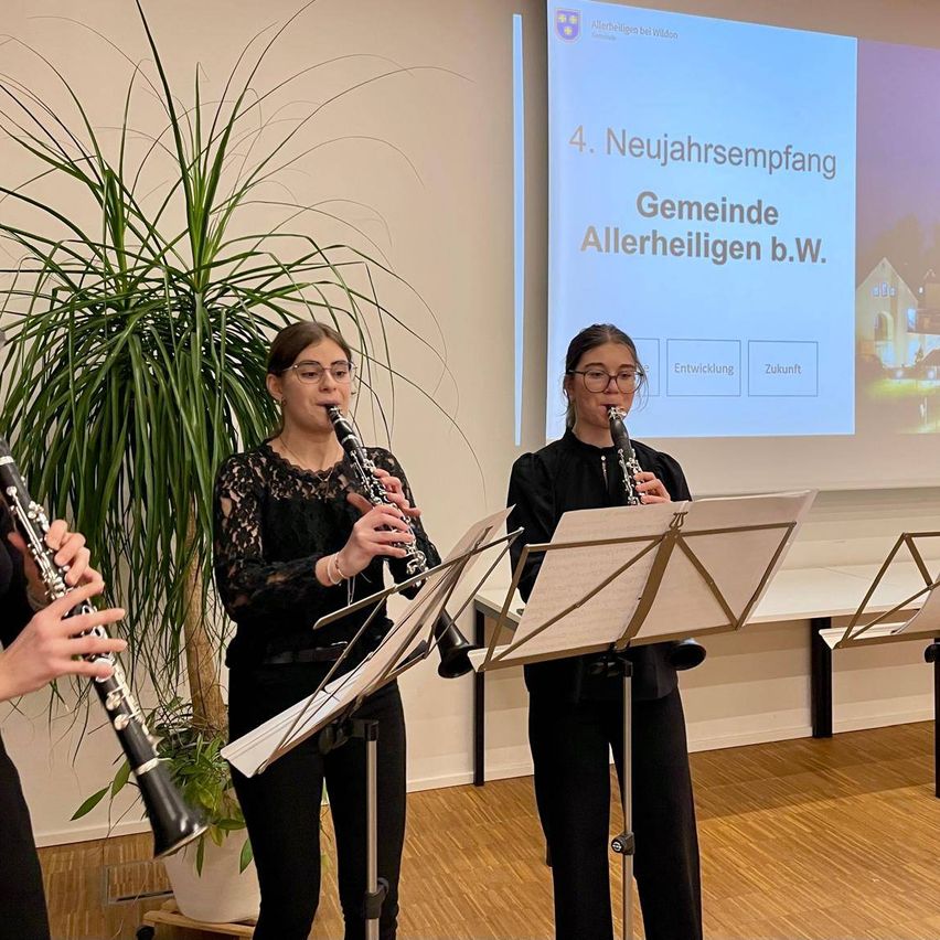 Drei Frauen spielen Klarinetten in einem Raum, in dem eine Beamerleinwand mit dem Text '4. Neujahrsempfang Gemeinde Allerheiligen b.W.' hinter ihnen steht. Sie stehen vor Notenständern.