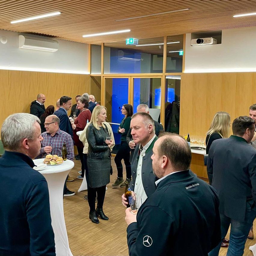 Eine Gruppe von Menschen versammelt sich in einem Raum, einige halten Getränke, andere unterhalten sich. Ein Mann hält eine Flasche und einen Becher. Ein anderer Mann hält einen Teller mit Sandwiches. Eine Frau steht in der Nähe eines Tisches mit Essen.