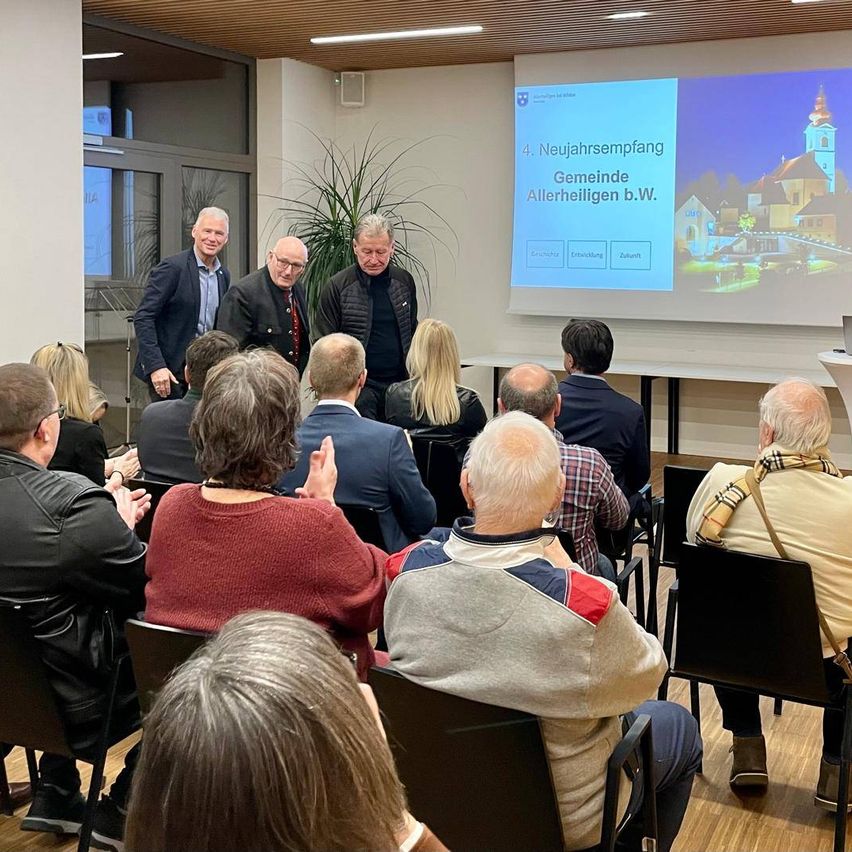 Mehrere Menschen sind drinnen versammelt und sitzen in Stühlen vor einer Präsentation auf einer Leinwand. Vier Männer stehen vor der Menge und scheinen zu sprechen. Eine Pflanze steht in der Ecke in der Nähe der Tür.