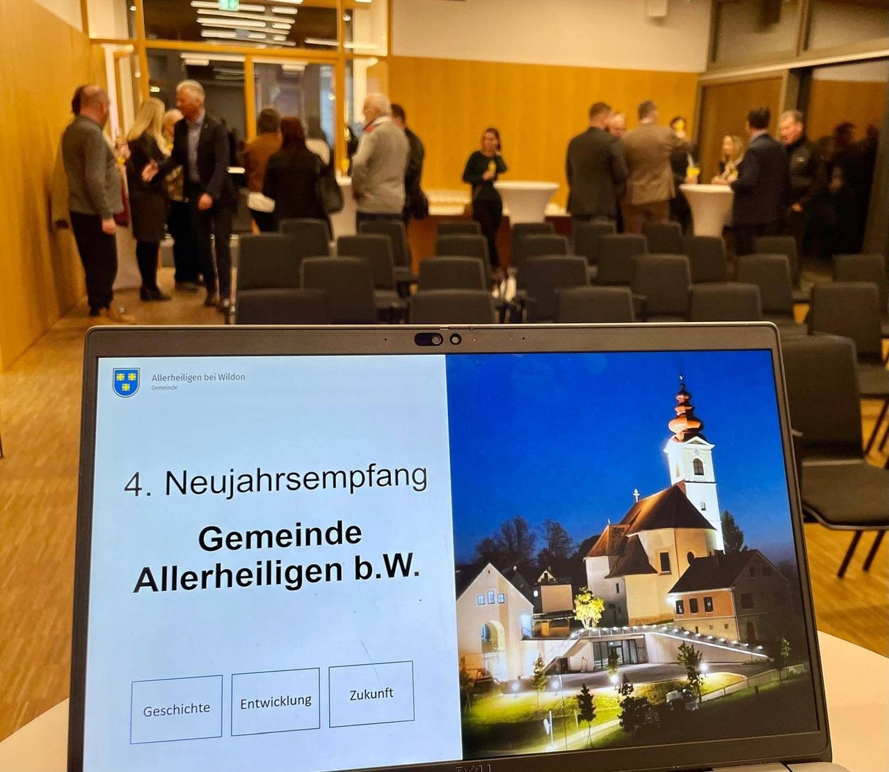 Ein Monitor zeigt eine Präsentation mit dem Titel '4. Neujahrsempfang Gemeinde Allerheiligen b.W.'. Im Hintergrund stehen und unterhalten sich Personen.