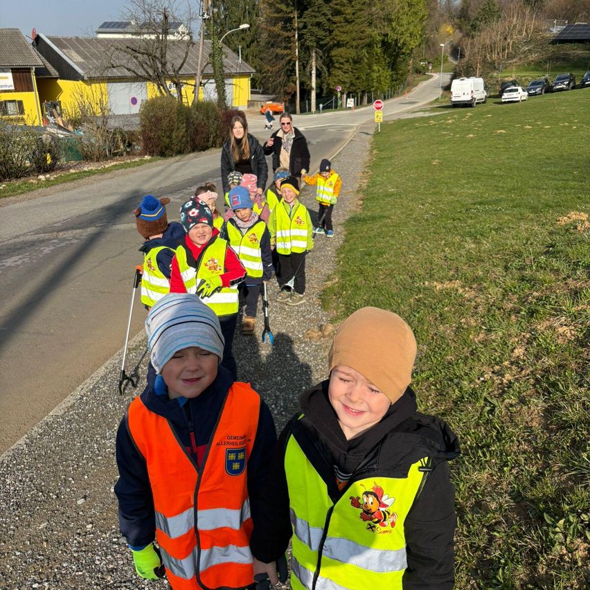 Eine Gruppe von Kindern und Erwachsenen geht mit Warnwesten auf einer Straße. Sie sind in der Nähe eines Grasbereichs mit gelben Gebäuden im Hintergrund.