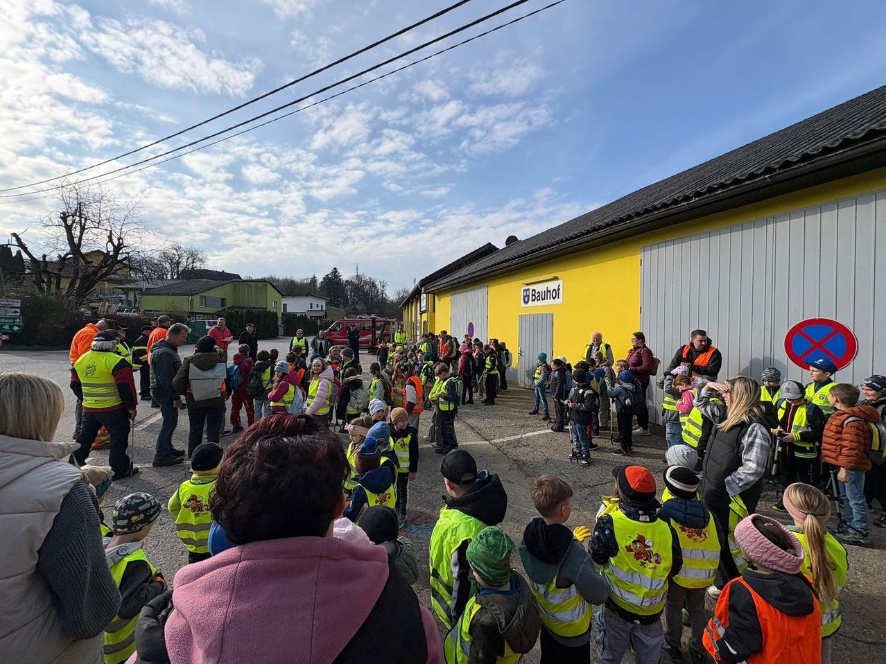 Eine Gruppe von Kindern in gelben Westen steht vor einem Gebäude. Einige Erwachsene sind ebenfalls anwesend, und ein Feuerwehrwagen ist im Hintergrund zu sehen.
