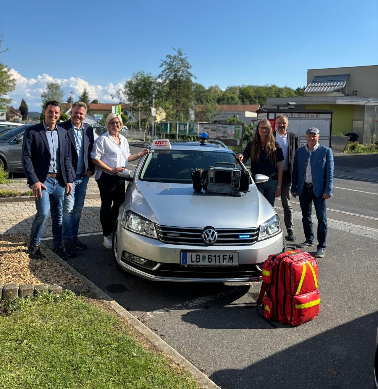 Eine Gruppe von Menschen steht neben einem silbernen Auto mit einem Schild obenauf. Sie alle lächeln. Das Auto hat ein Nummernschild. In der Nähe steht ein roter Rucksack mit gelben und weißen Markierungen.