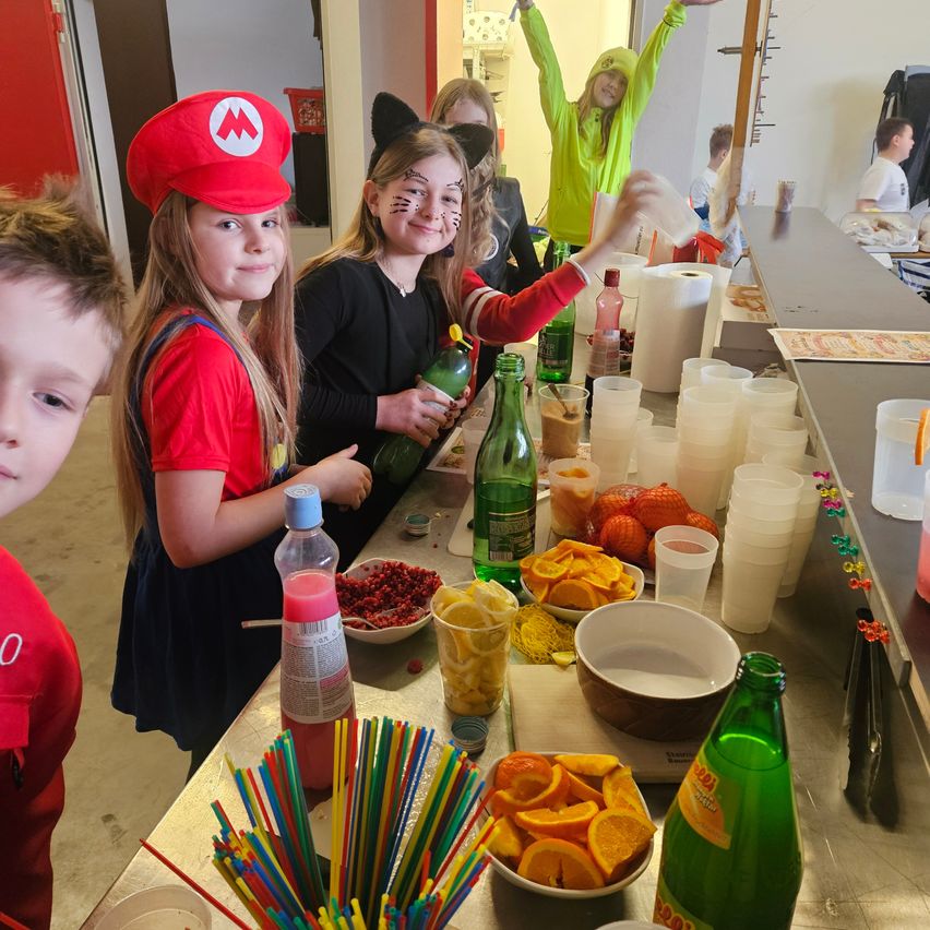 Kinder in Kostümen stehen hinter einem Tresen voller Getränke und Snacks. Ein Mädchen hat eine Mario-Mütze und ein anderes hat Gesichtsbemalung.