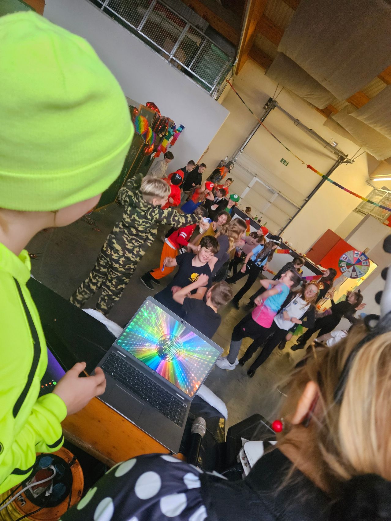 Eine Gruppe von Kindern in bunten Kostümen tanzt in einem Raum mit einem Laptop und einem DJ-Pult im Vordergrund.