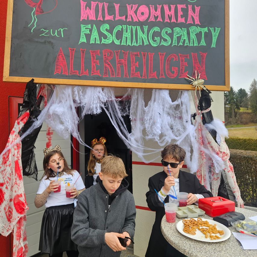Vier Kinder stehen vor einem Gebäudeeingang, wahrscheinlich für eine Halloween-Party. Ein Kreidetafel über ihnen steht 'Willkommen zur Faschingsparty Allerheiligen'. Die Kinder sind in Kostümen gekleidet, und eines von ihnen trinkt aus einem Becher. Ein Tisch in der Nähe hat Essen und Getränke.