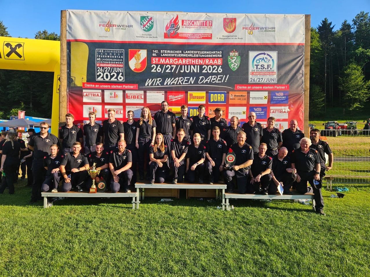 Eine Gruppe von Feuerwehrleuten aus verschiedenen Bezirken posiert für ein Foto mit einem großen Banner hinter ihnen. Sie stehen auf einem Rasenfeld.