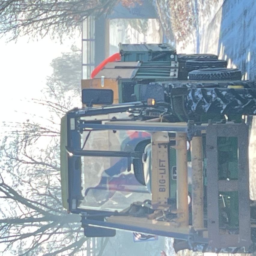 Ein großer grüner Traktor mit der Aufschrift 'BIG-LIFT' an der Seite ist in einem verschneiten Bereich geparkt. Bäume und ein unscharfer Hintergrund sind im Bild zu sehen.