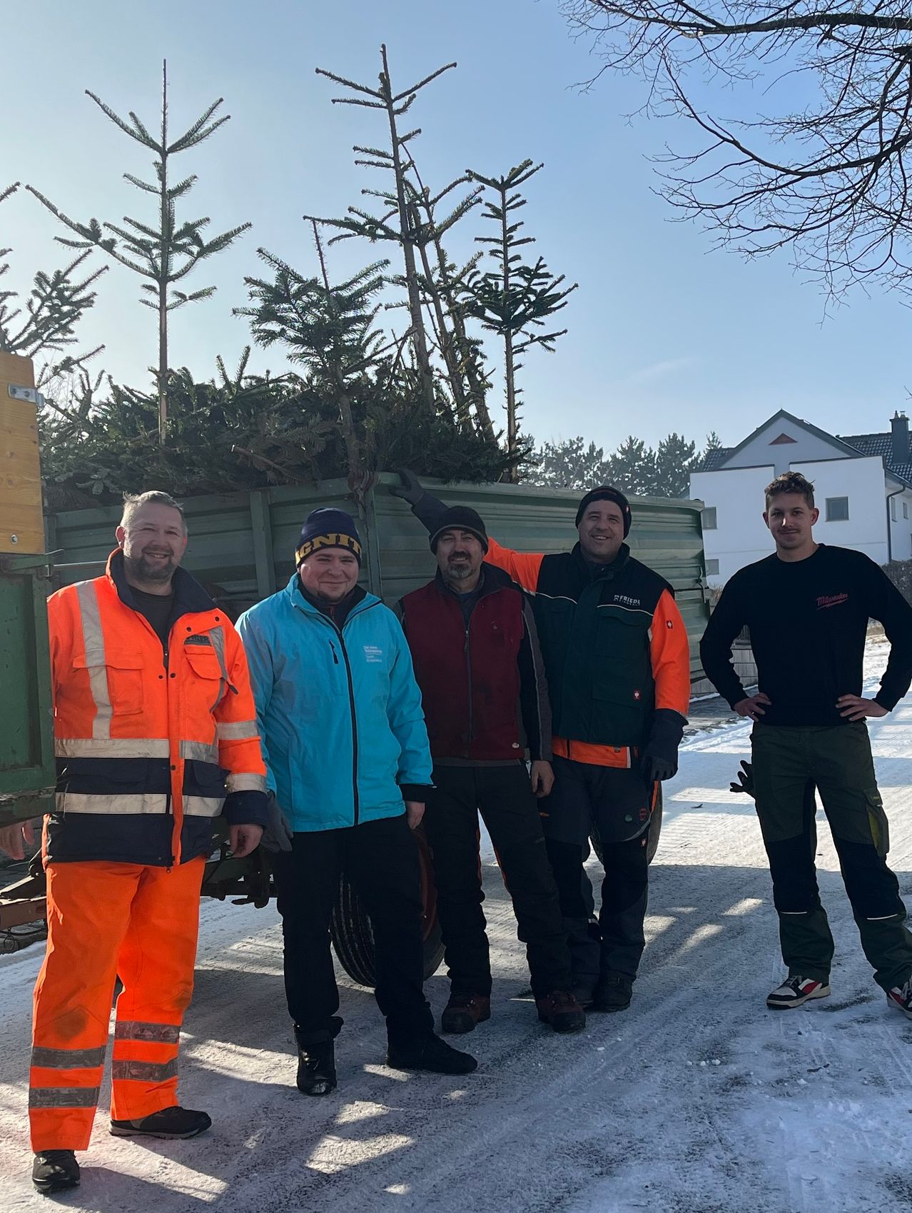 Fünf Männer in Winterkleidung stehen vor einem mit Tannenbäumen gefüllten Lastwagen, wahrscheinlich für ein Foto posierend. Dahinter ist ein weißes Haus mit Fenstern zu sehen.
