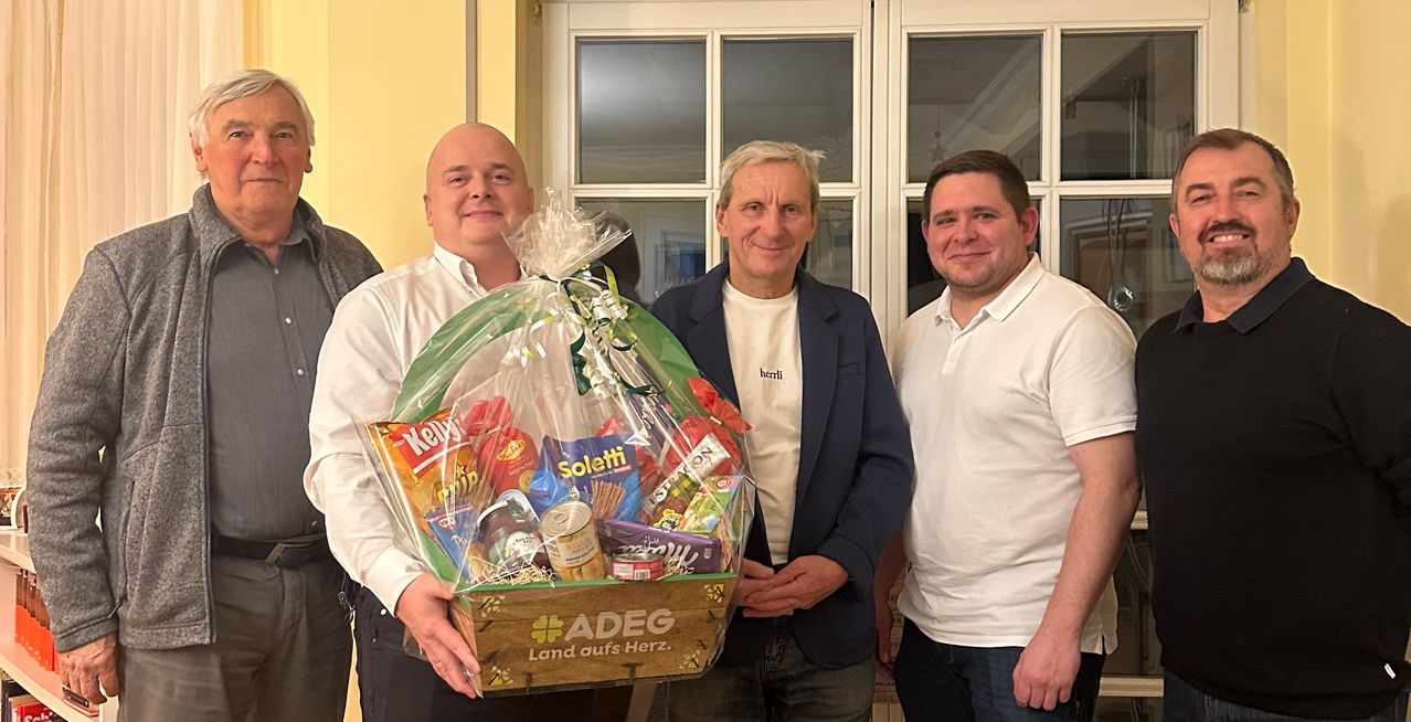 Drei Männer stehen in einem Raum mit einem großen Geschenkkorb, gefüllt mit Snacks. Der Mann auf der linken Seite hält den Korb und lächelt. Der Mann in der Mitte trägt einen Mantel und sieht glücklich aus. Der Mann auf der rechten Seite trägt ein Poloshirt und lächelt ebenfalls.