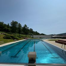 Ein Freibad mit klarem blauem Wasser und schwarzen Bahnlinien. Bäume und Grünflächen umgeben das Becken, mit Bänken und einer weißen Wand im Hintergrund.