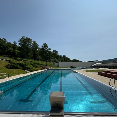 Ein Freibad mit klarem blauem Wasser und schwarzen Bahnlinien. Bäume und Grünflächen umgeben das Becken, mit Bänken und einer weißen Wand im Hintergrund.