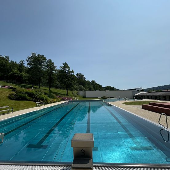 Ein Freibad mit klarem blauem Wasser und schwarzen Bahnlinien. Bäume und Grünflächen umgeben das Becken, mit Bänken und einer weißen Wand im Hintergrund.