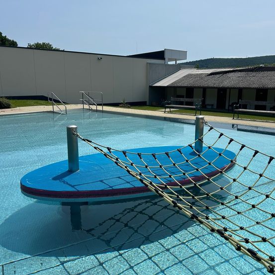 Ein leerer Swimmingpool mit einer blauen Plattform und einem Netz in der Mitte. Es gibt zwei Bänke an der Seite und ein Gebäude im Hintergrund.