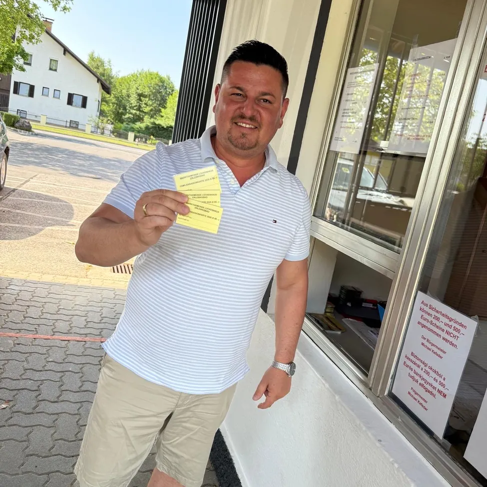 Ein Mann hält gelbe Tickets vor einer Glastür. Er trägt ein gestreiftes Hemd und khakifarbene Shorts. Hinter ihm steht ein Gebäude mit einem geparkten Auto und einem weißen Haus im Hintergrund.