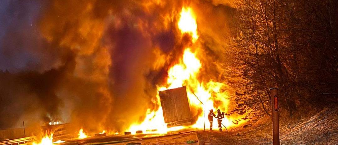 Ein brennendes Fahrzeug steht am Straßenrand, während zwei Feuerwehrleute die Flammen bekämpfen. Die Szene spielt sich bei Nacht ab, wobei Rauch und Feuer auf die umliegenden Bäume übergreifen.