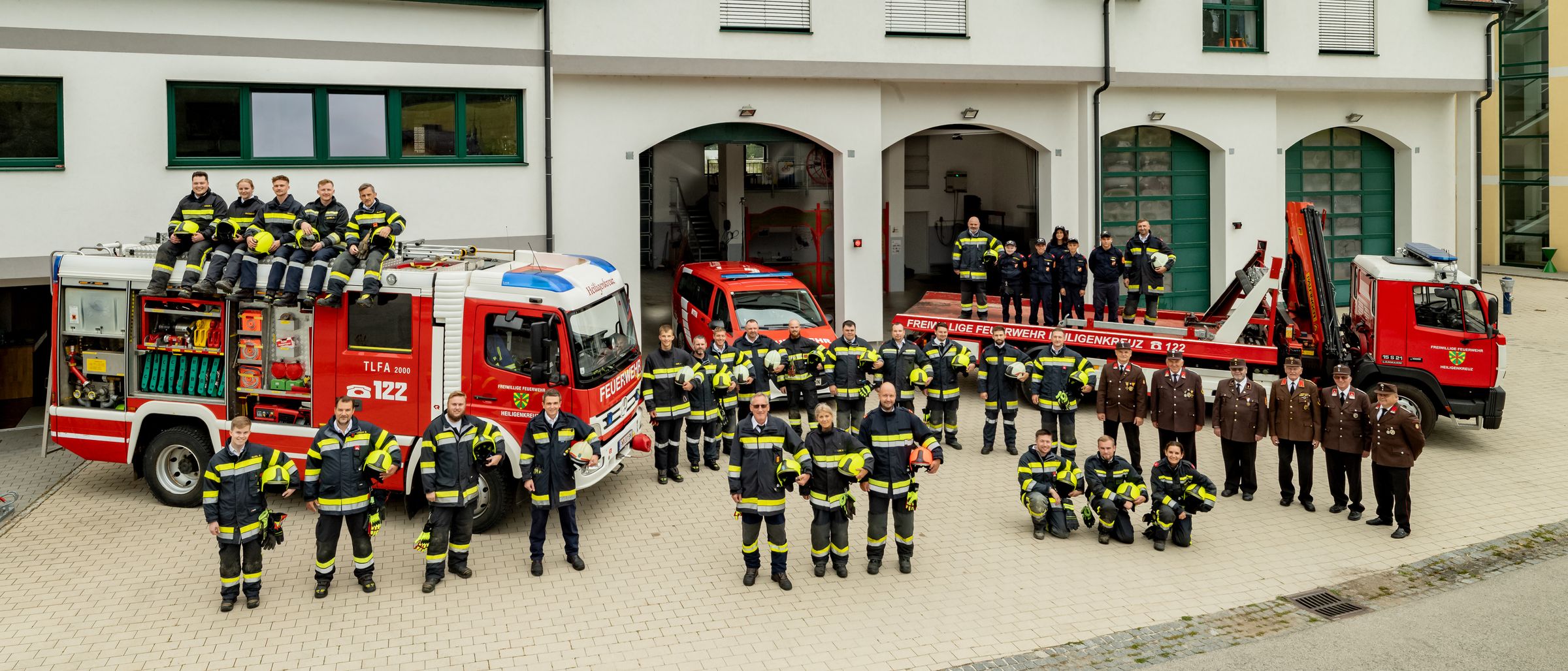 Eine Gruppe von Feuerwehrleuten, einige stehend und andere kniend, posiert vor einem Feuerwehrhaus. Sie tragen gelbe und schwarze Uniformen und Helme. Im Hintergrund sind zwei Feuerwehrwagen geparkt.